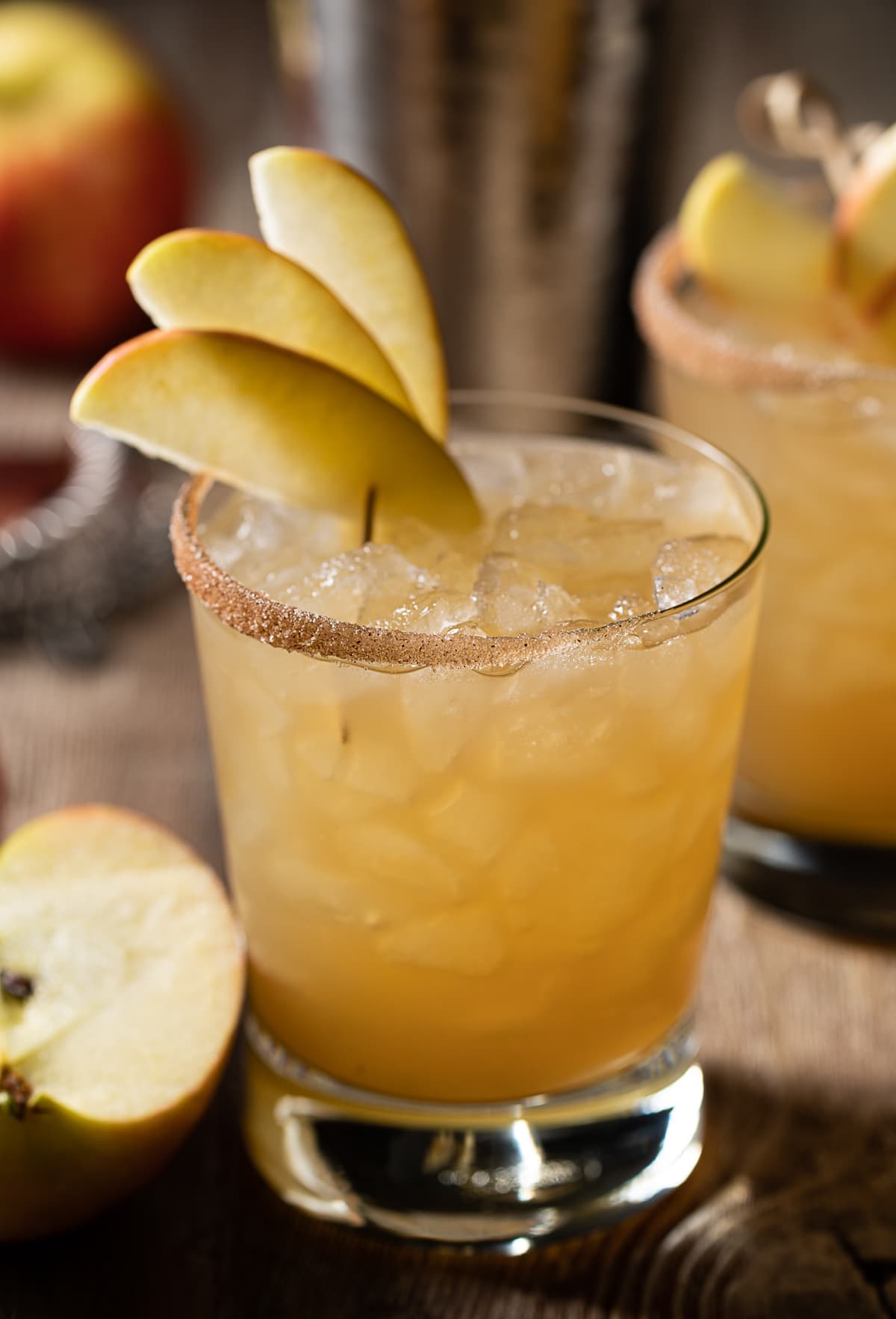 gold colored alcohol mixture in rocks glasses filled with ice, thin apple slices, cocktail shaker, half an apple