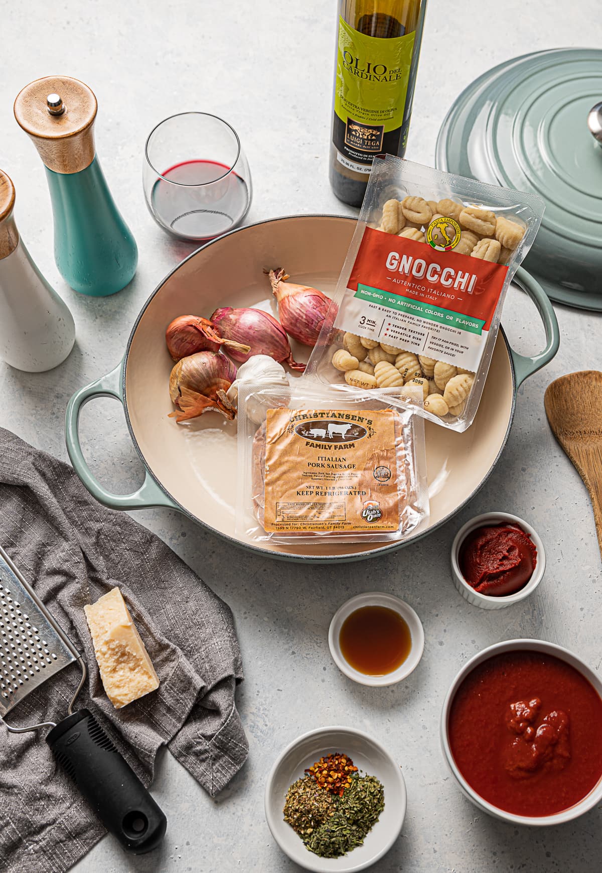 large aqua pan full of recipe ingredients, package of Italian sausage, package of gnocchi, shallots, garlic, bowls of tomato sauce, bottle of olive oil, chink of parmesan cheese, glass of red wine