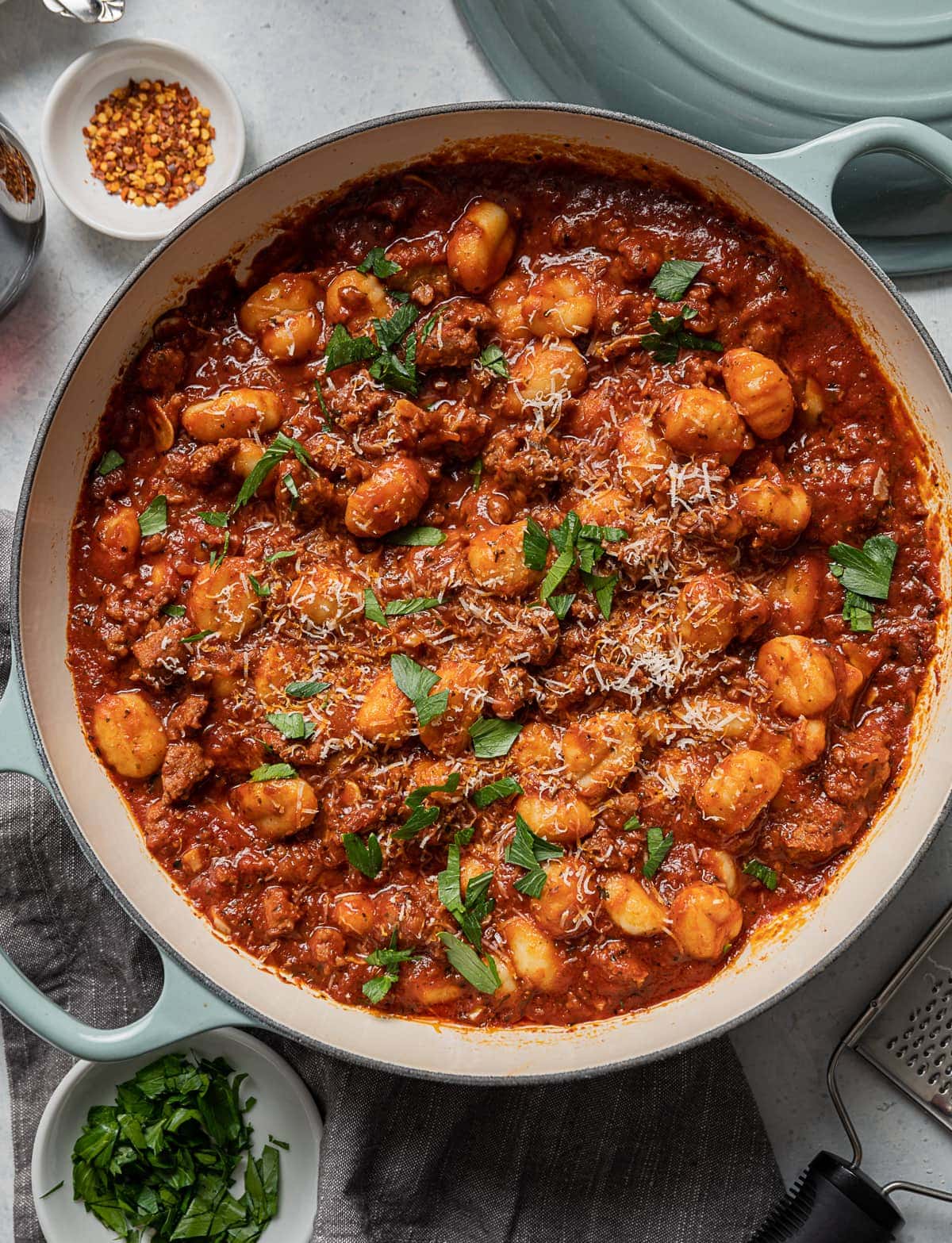 large aqua pan with sausage and gnocchi with red tomato sauce, bowl of green parsley, bowl of red pepper flakes, grater 