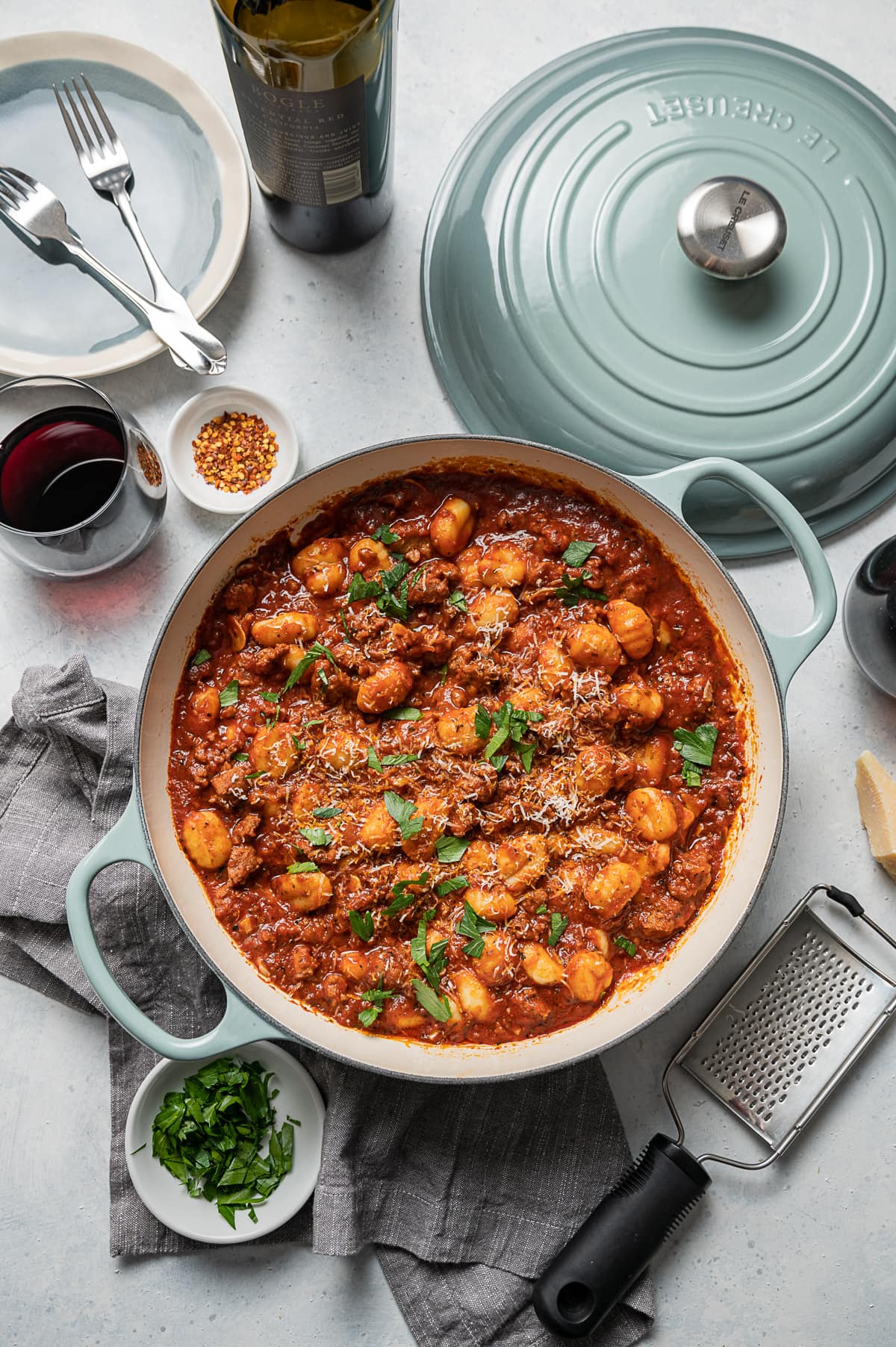 large aqua pan with sausage and gnocchi with red tomato sauce, glasses of red wine, bowl of green parsley, bowl of red pepper flakes, plates, forks, bottle of wine