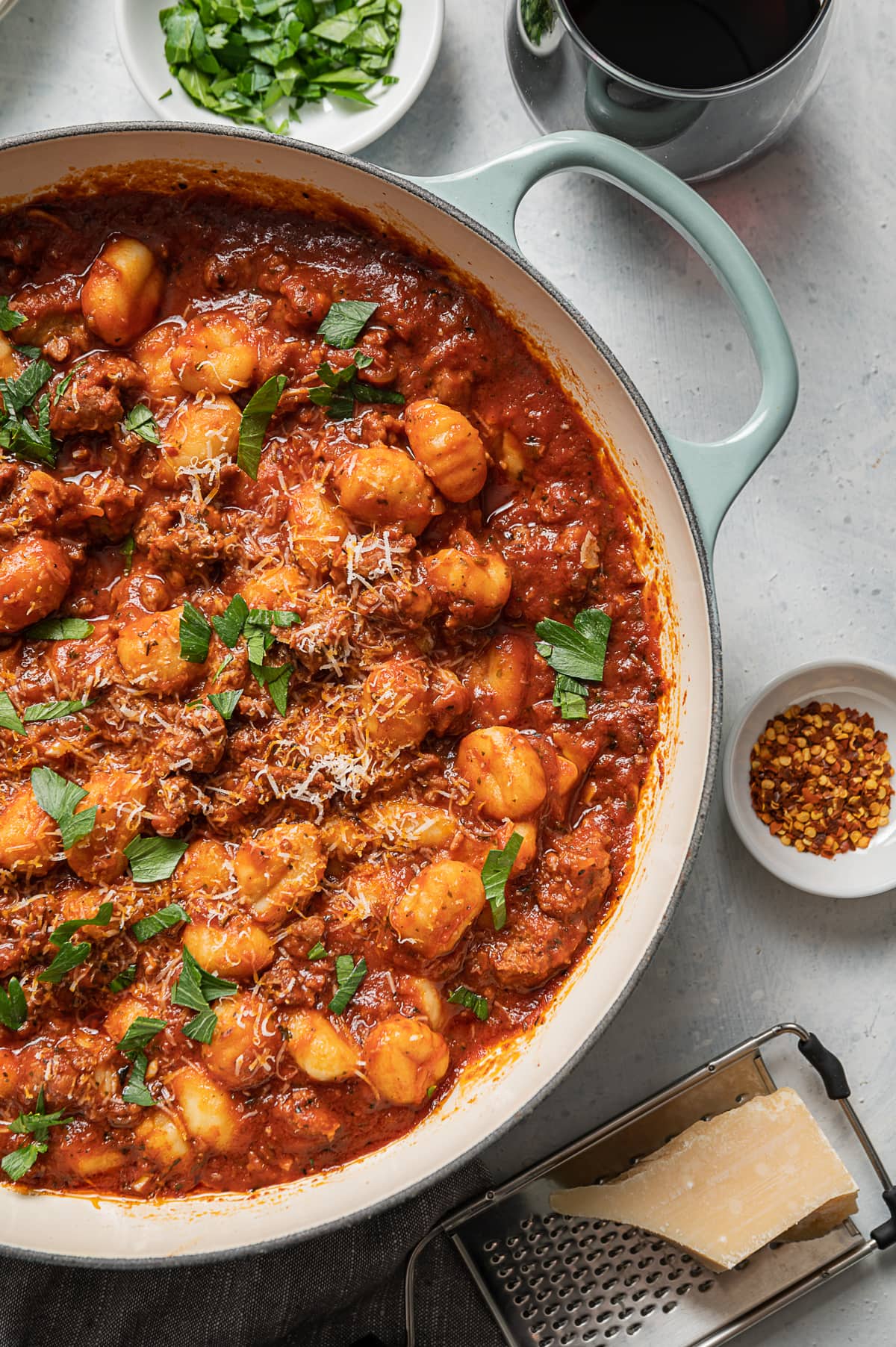 large aqua pan with sausage and gnocchi with red tomato sauce, glasse of red wine, bowl of green parsley, bowl of red pepper flakes, grater and chunk of parmesan cheese