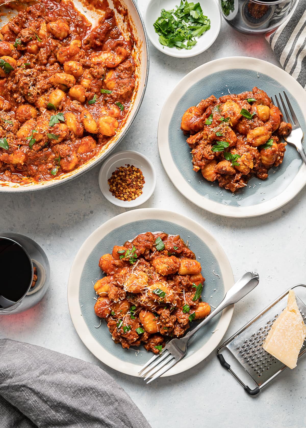 two small plates with red tomato sauce sausage and gnocchi, forks, glasses of wine, small bowls of red pepper flakes and fresh parsley, large bowl of red sauce with sausage and gnocchi