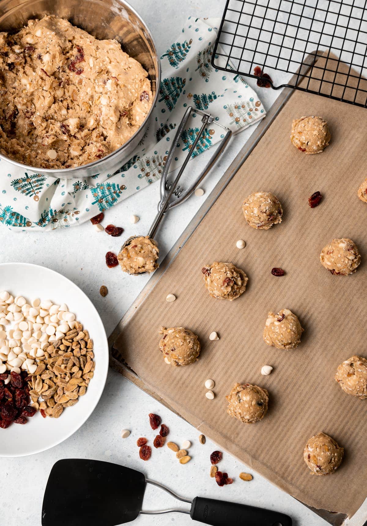 oatmeal cookie dough on a tray some dough in a stainless bowl some dough in a cookie scoop bowl of white chocolate chips cranberries and pistachios