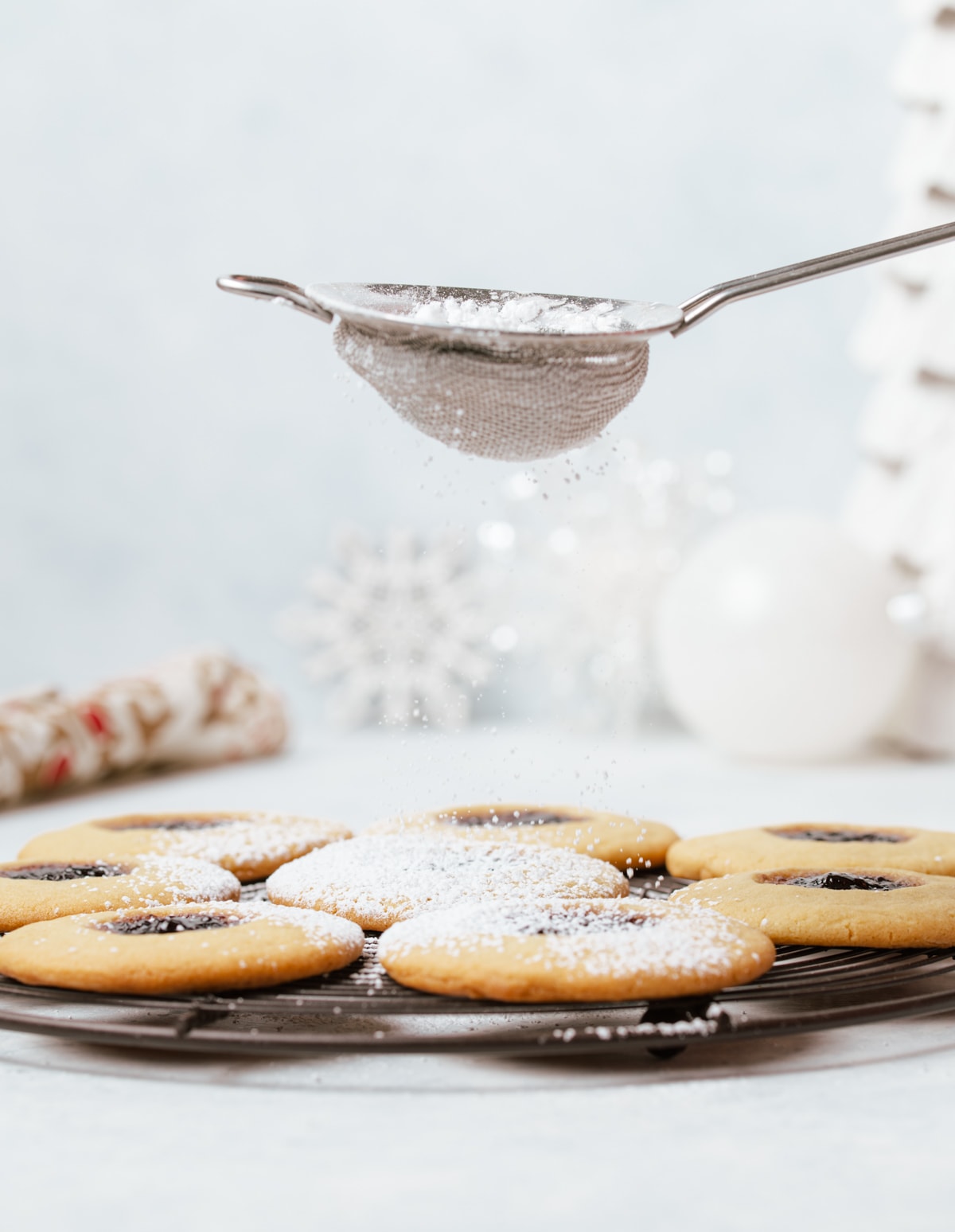 powdered sugar being dusted on round sandwich cookies with dark purple jam layered on a wire rack