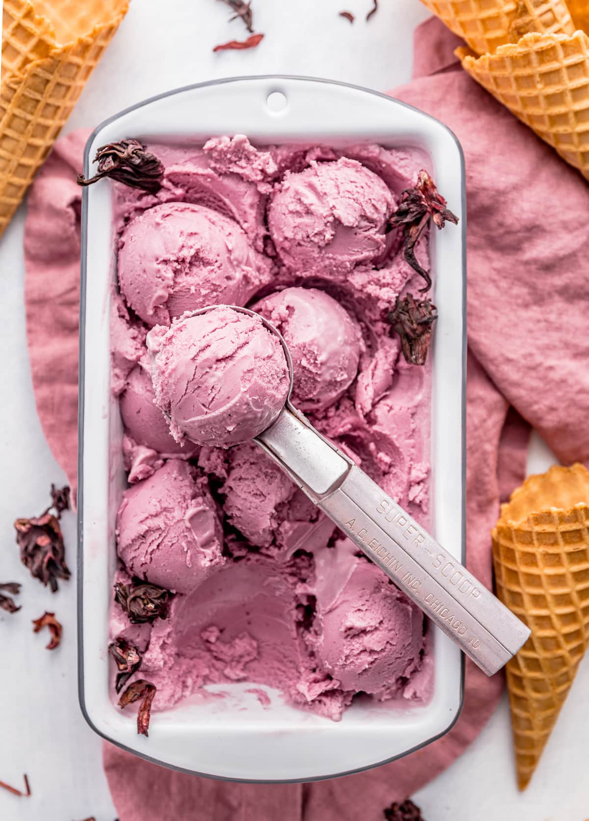 pink vegan coconut hibiscus ice cream in a white loaf pan with pink ice cream scoop and waffle ice cream cones