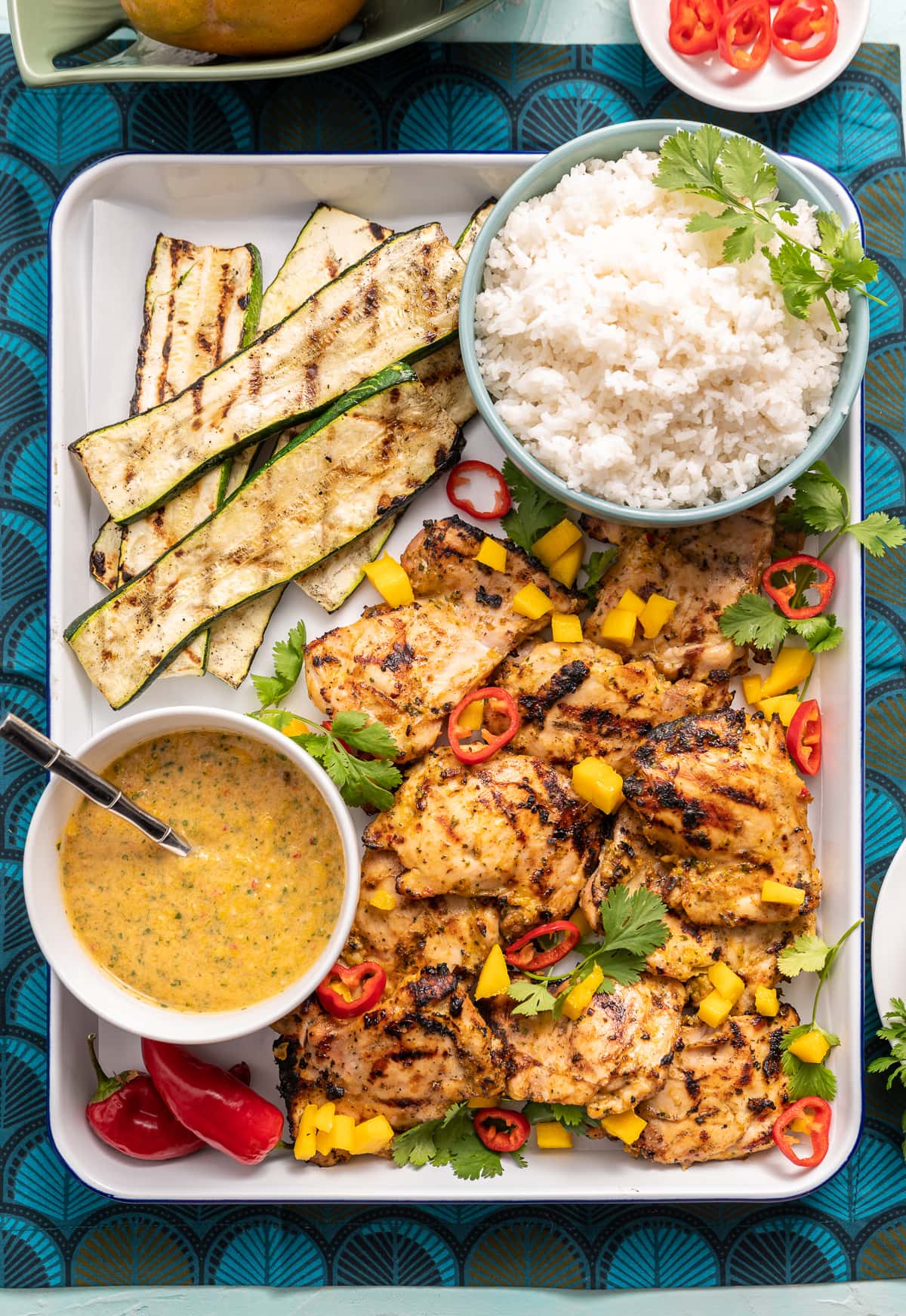 grilled chicken on a tray with mango and red chili slices cilanto pieces bowl of white rice bowl of yellow mango sauce grilled zucchini slices