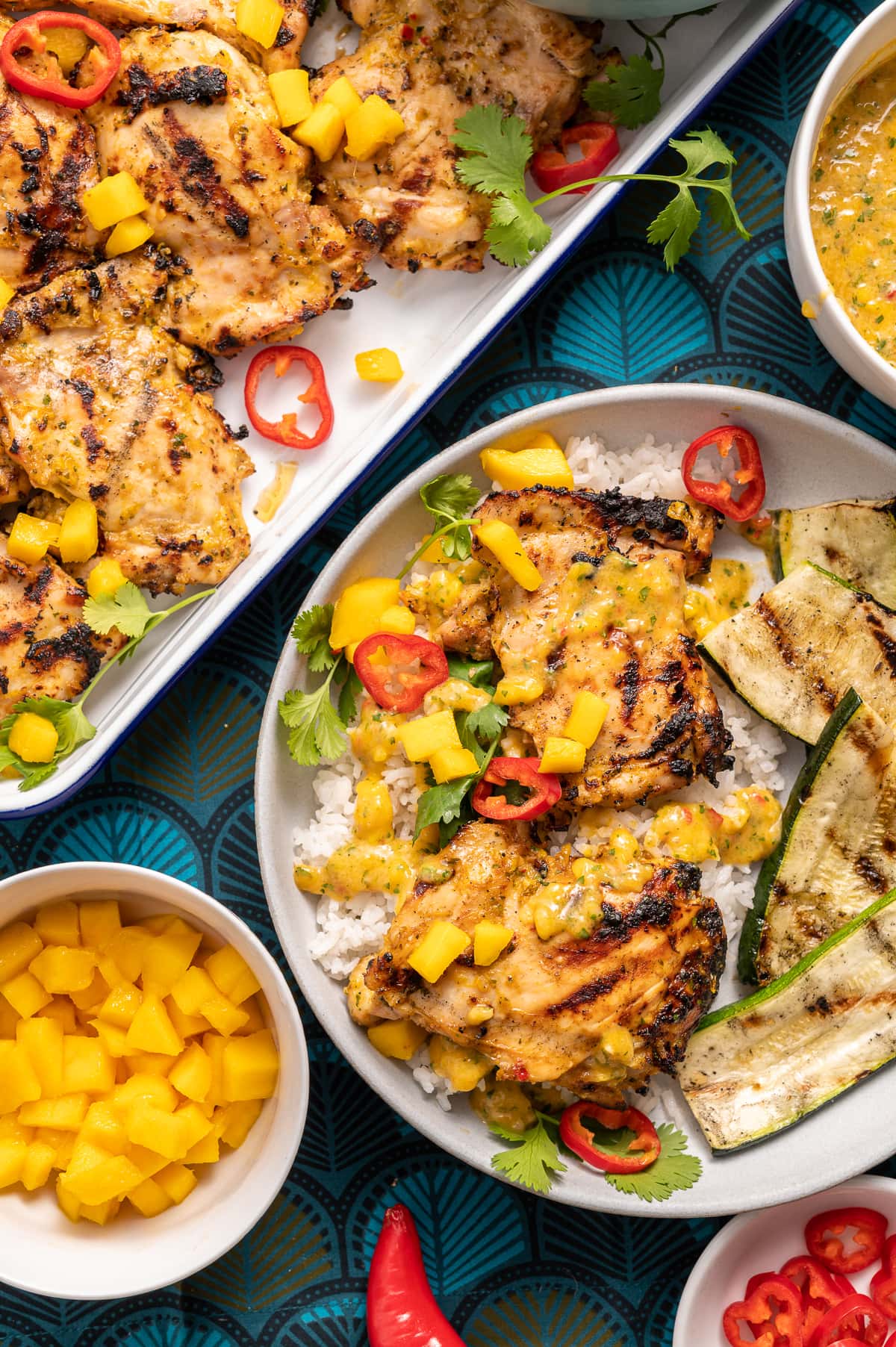 grilled chicken on a plate with mango and red chili slices cilanto pieces bowl of white rice bowl of yellow mango sauce grilled zucchini slices