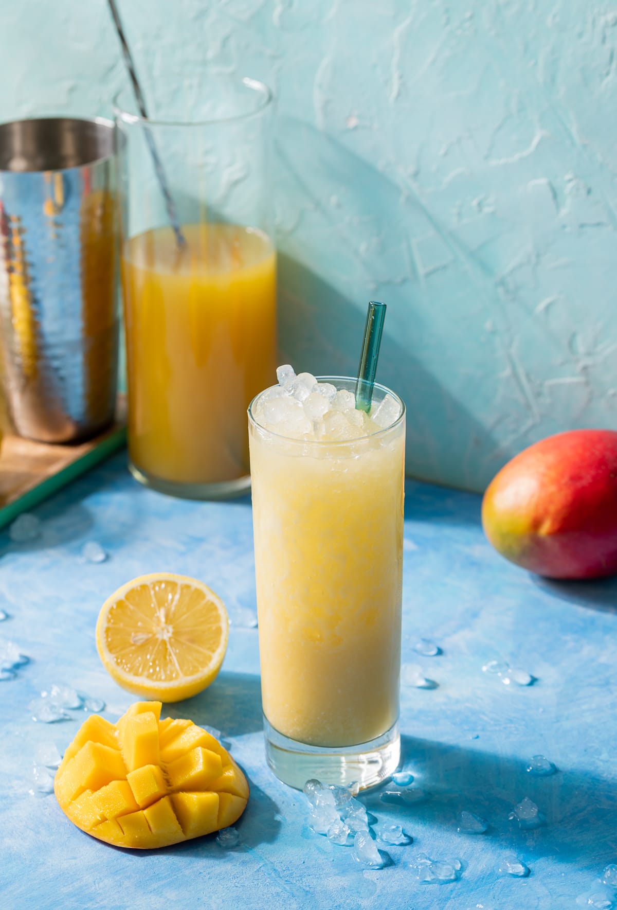 Shaken Coconut Mango Lemonade in a glass with mango and lemon slices tall glass of lemonade metal cocktail shaker