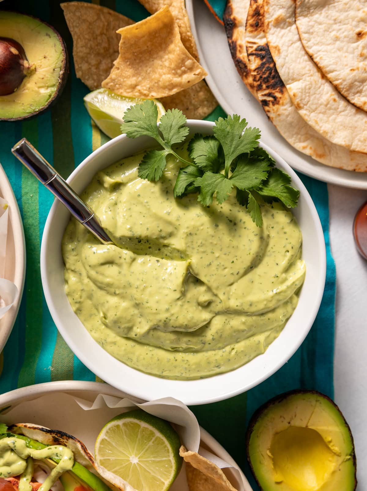 green lime avocado crema sauce in white bowl with spoon and fresh cilantro plate of tortillas chips avocado cut in half