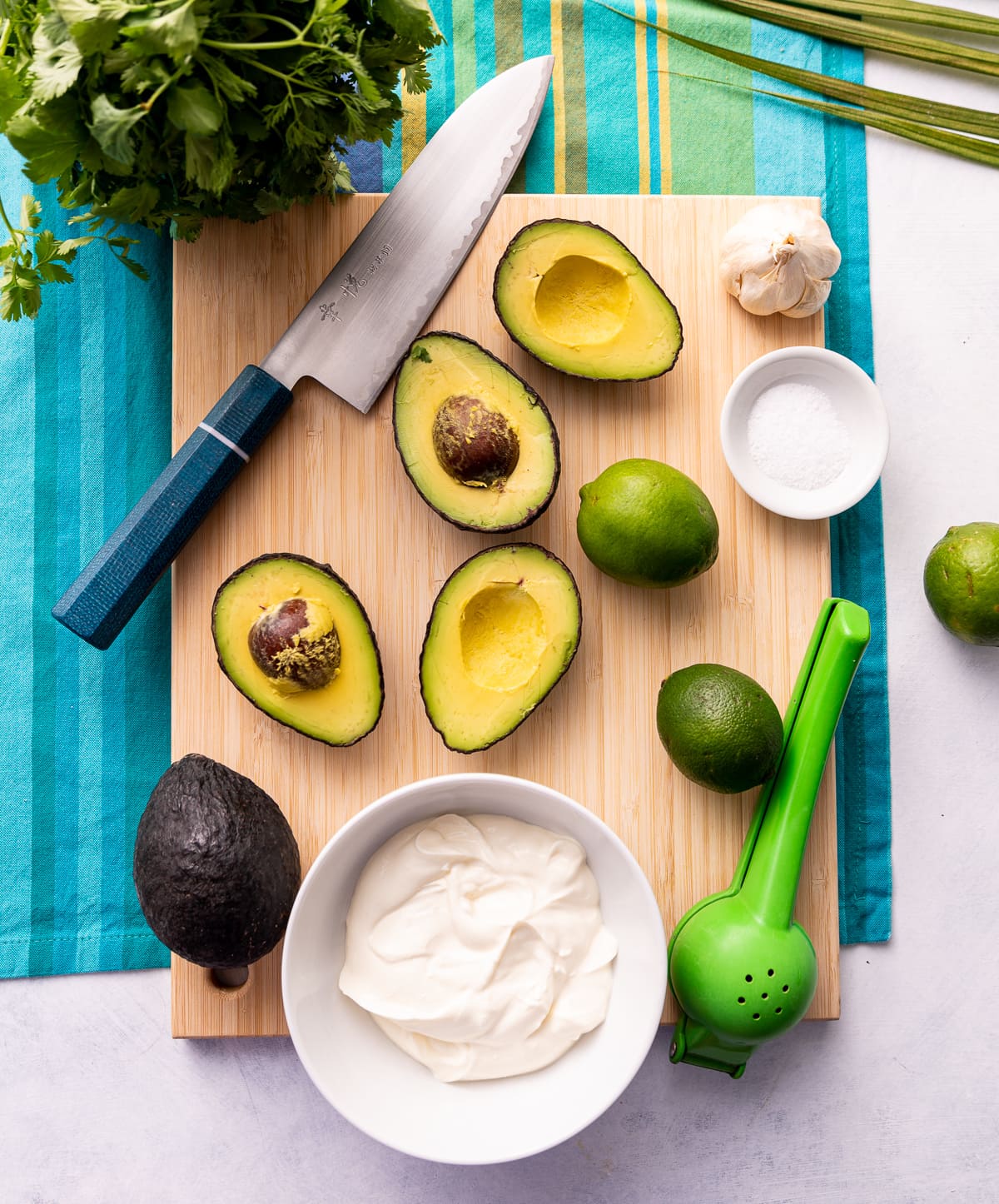 cutting board with avocados sliced in half whole limes knife with blue handle bowl of white sour cream green citrus press