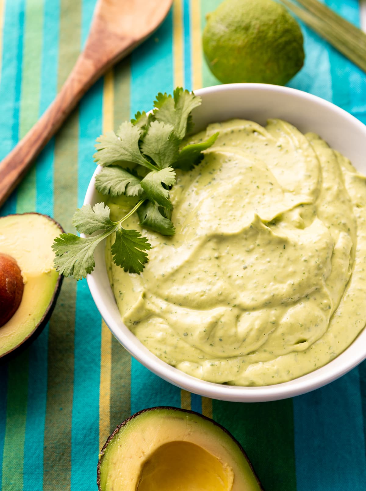 green lime avocado crema sauce in a white bowl with fresh green cilantro avocado cut in half blue striped napkin