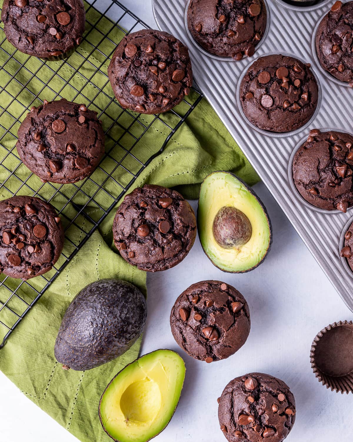 chocolate avocado muffins on a wire cooling rack and in a baking pan with cut avocados and a green napkin