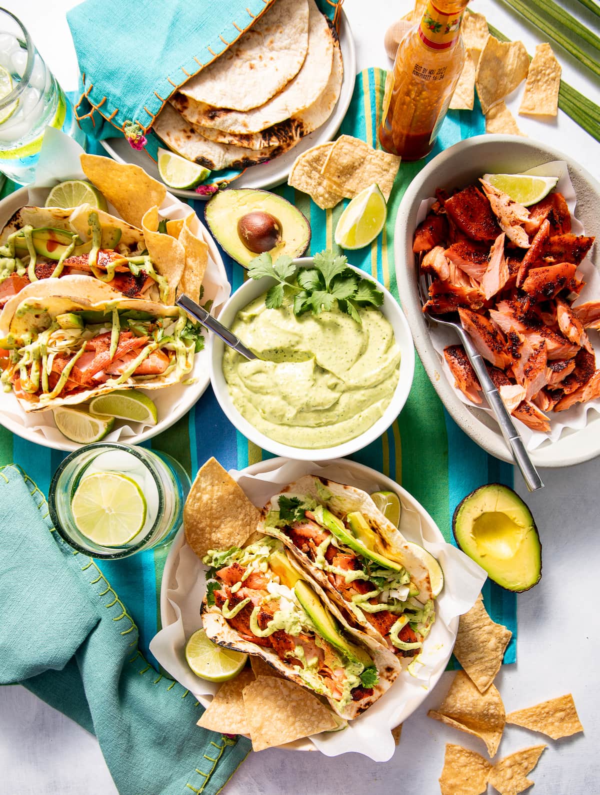 whole table spread of bowls of smoked salmon tacos with a bowl of green avocado crema bowl of smoked salmon bottle of hot sauce plate of tortillas chips and avocado halves
