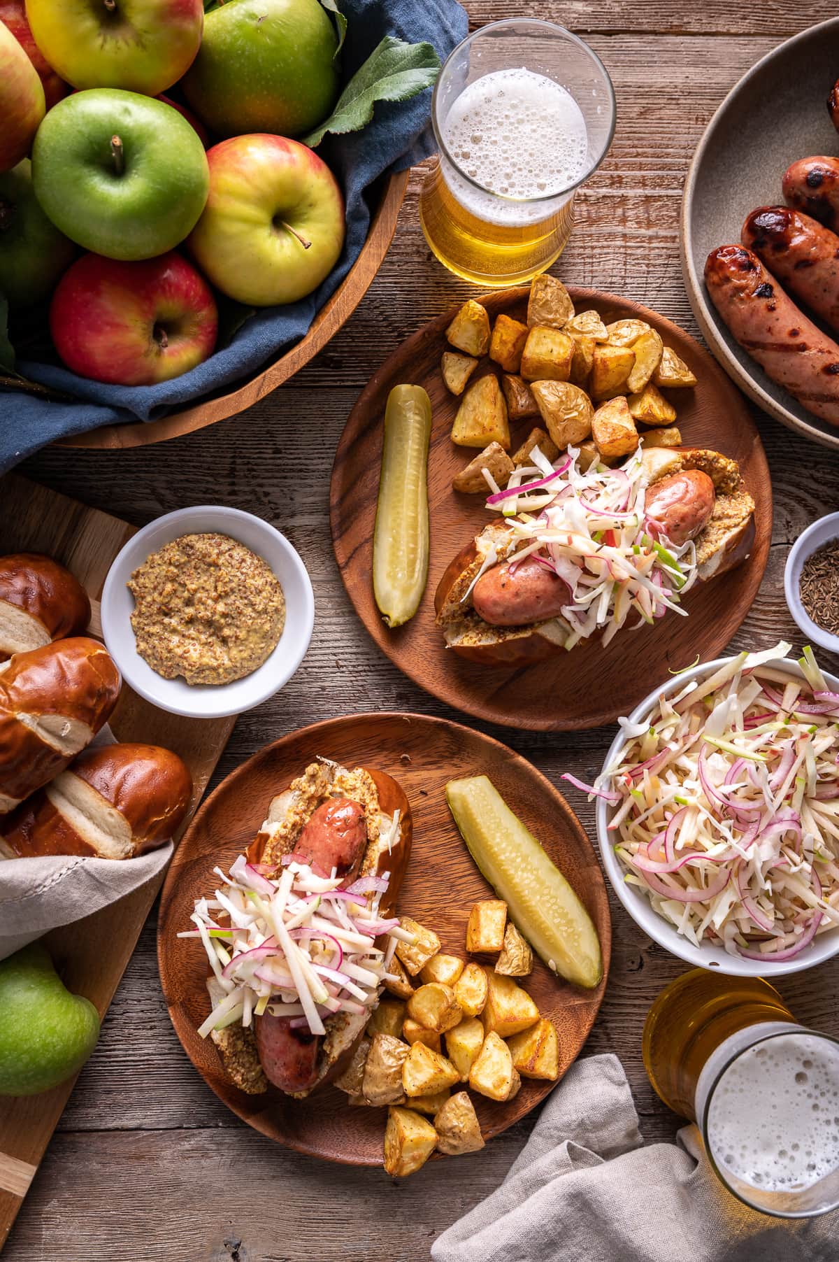 sausages on pretzel rolls on brown plates with apple slaw pickles roast potatoes bowl of apples glasses of beer bowl of german mustard