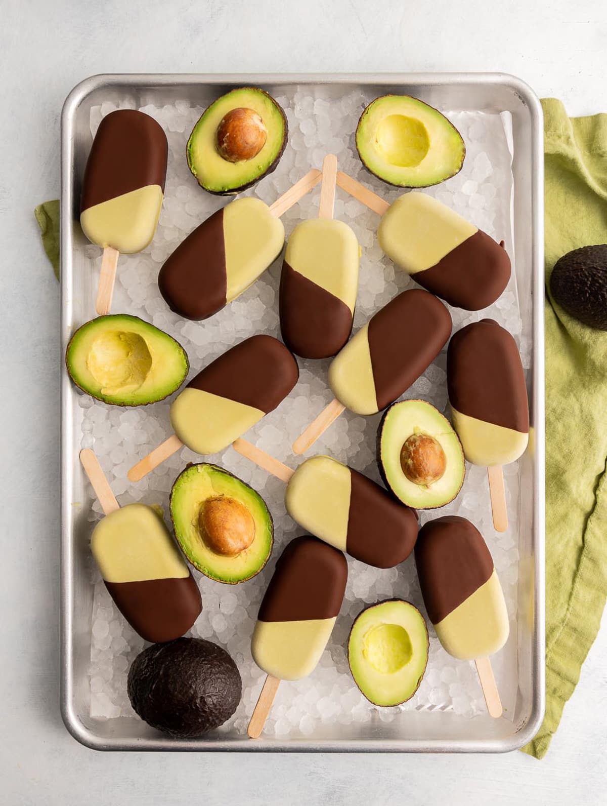 tray of ice with green avocado ice cream bars half dipped in chocolate on the ice with avocados cut in half
