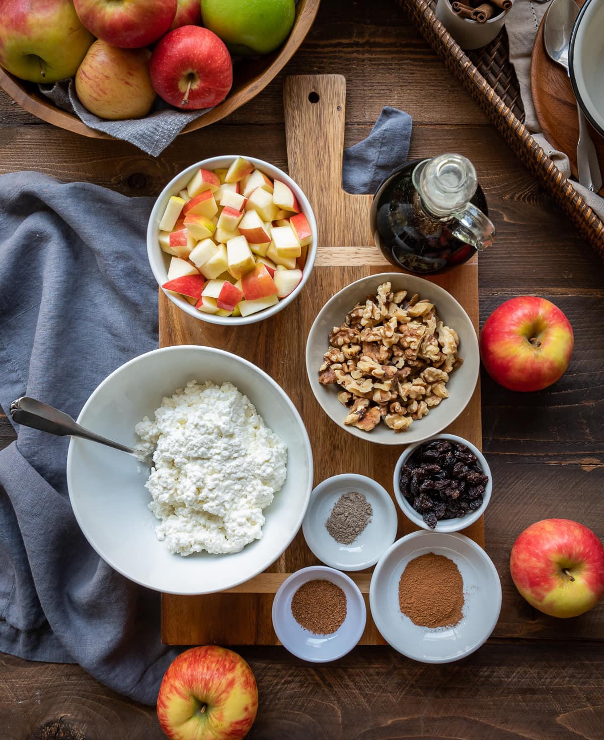 brown cutting board with bowls filled with ingredients for maple apple cinnamon cottage cheese breakfast bowls bowl of cottage cheese bowl of apples bowl of nuts small bowls of spices whole apples