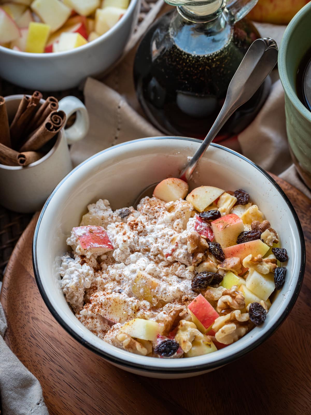 apple chunks walnuts raisins and spices mixed with cottage cheese in white bowl on brown plate small cup of whole cinnamon sticks