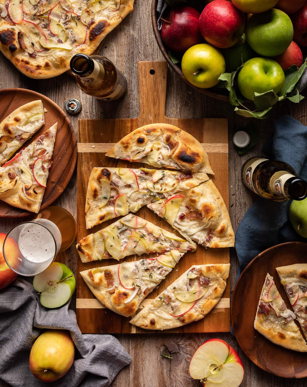 flammkuchen flatbread with apples onion and bacon cut into slices on on a cutting board with whole apples and bottles of german beer
