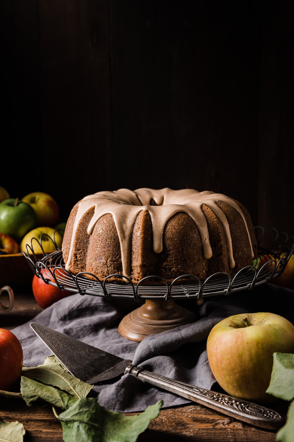 brown bundt cake with light brown icing on a wire cake stand with cake serving tool and whole yellow red and green apples