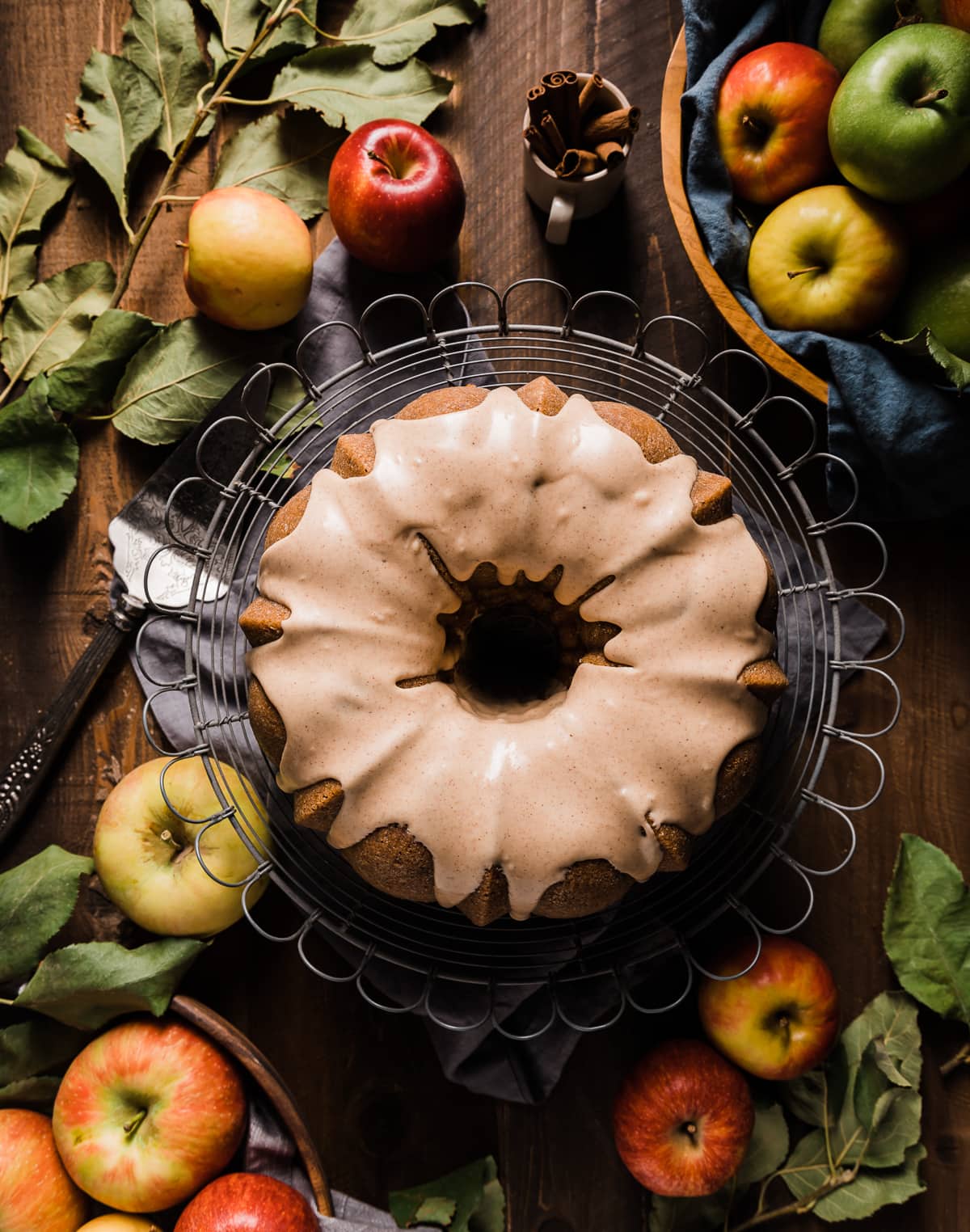 brown sugar bundt cake with cinnamon icing on a wire cake plate with whole yellow red and green apples grey napkin