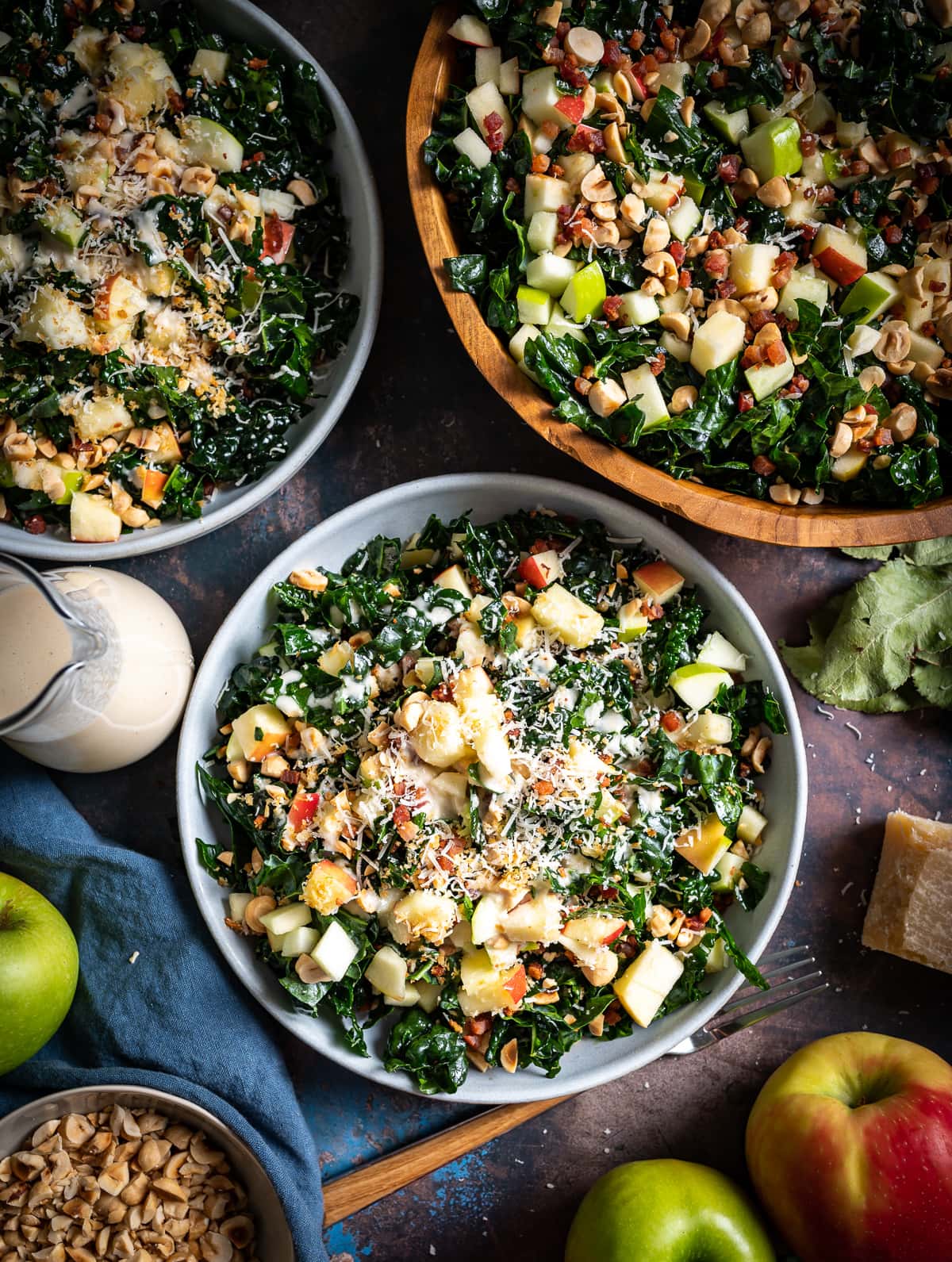large wood serving bowl and two smaller blue ceramic bowls filled with chopped tuscan kale with hazelnuts apples parmesan cheese bottle of vinaigrette