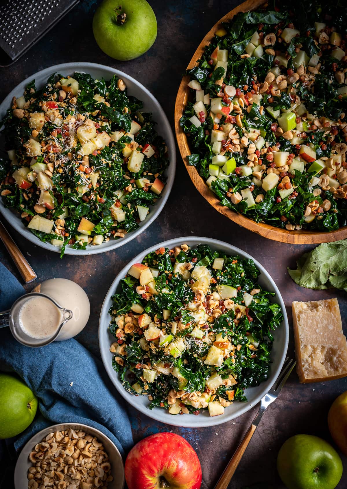 bowls of chopped kale salad with diced apples, hazelnuts, whole apples whole piece of parmesan cheese bottle of vinaigrette
