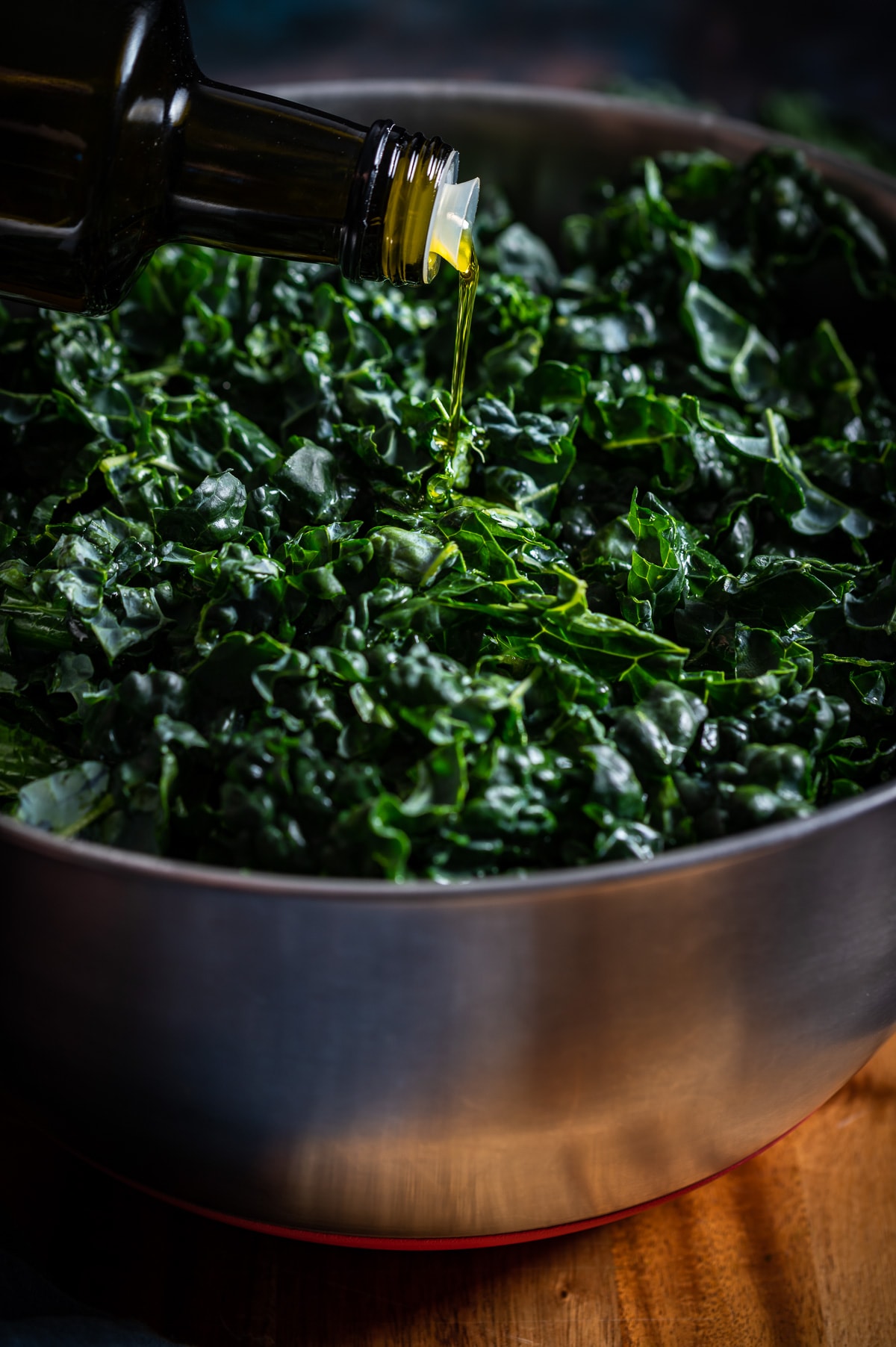 olive oil being poured into a large metal bowl of chopped tuscan kale