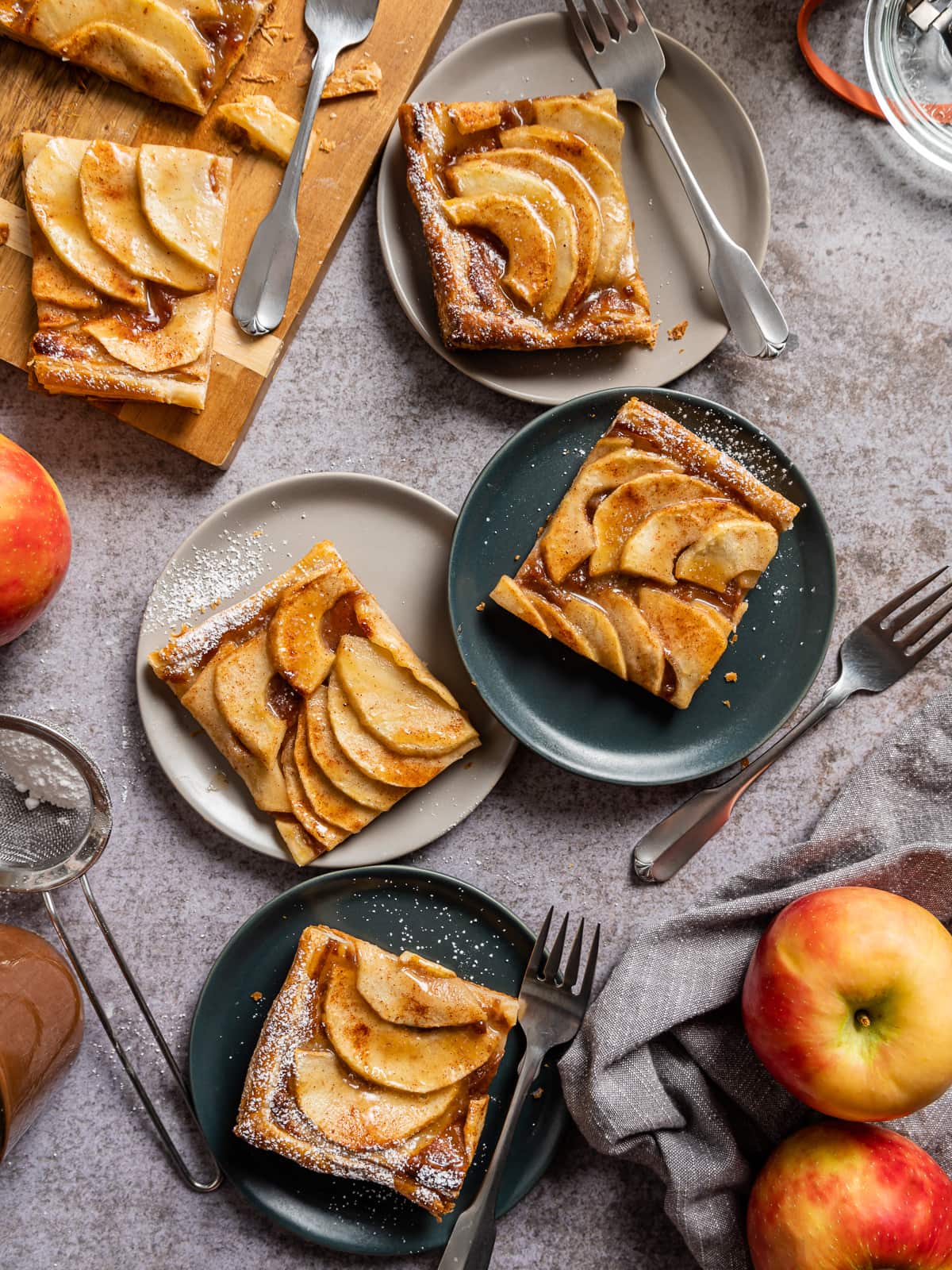 Square slices of apple puff pastry tarts on light gray and dark gray plates forks and whole apples