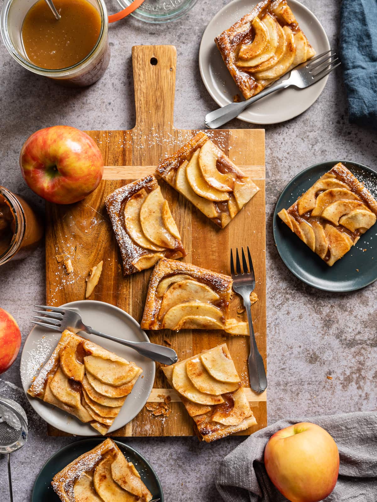 puff pastry apple tart slices on a cutting board with forks a few slices on light gray and dark gray plates whole red and yellow apples