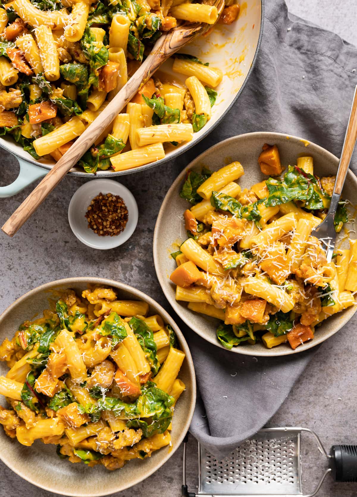 two beige bowls and a pale blue pan filled with tube shaped pasta with orange honeynut squash sauce and pieces green swiss chard pieces 