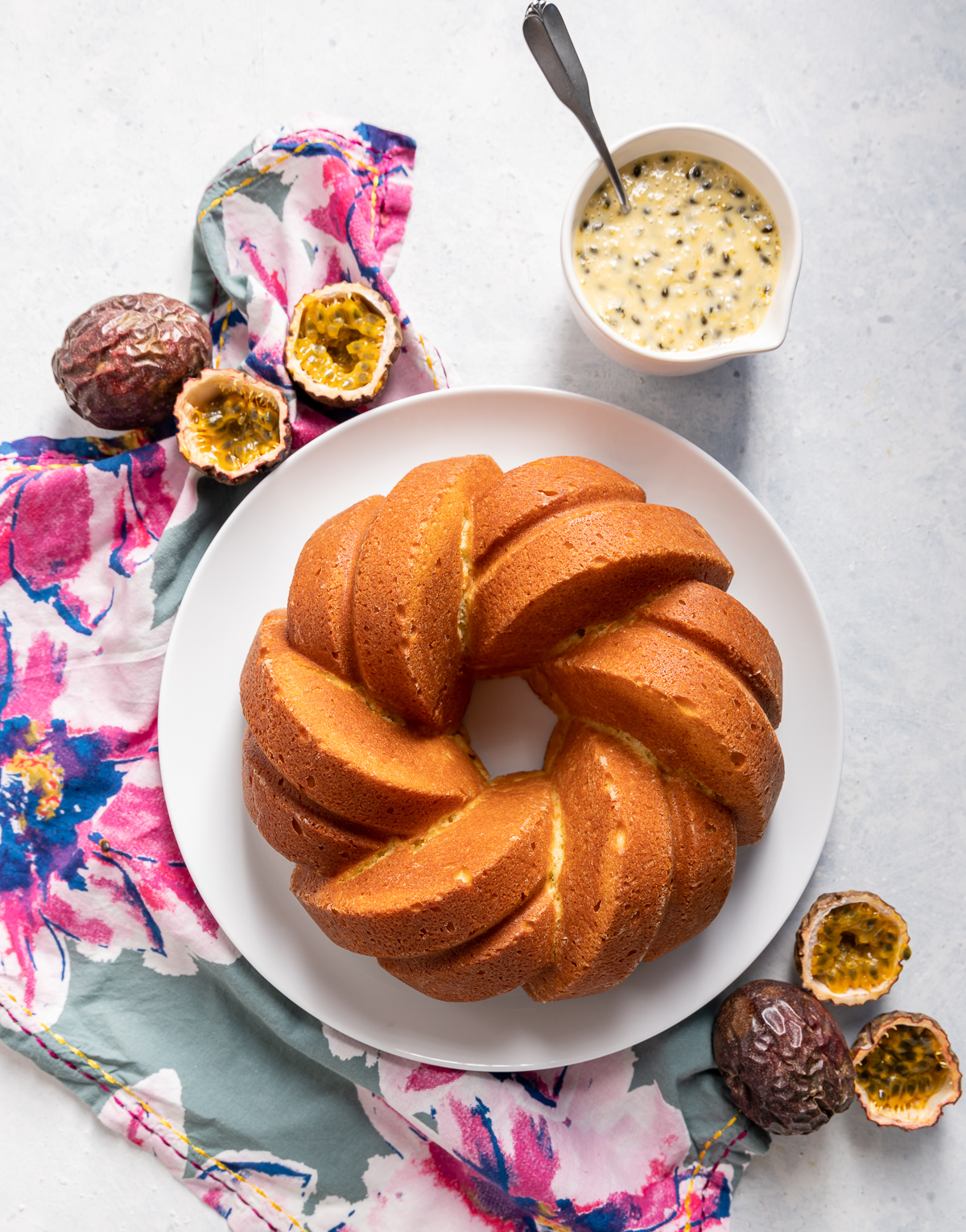 golden yellow braided bundt cake on a white plate blue and pink flower napkin whole and halved passionfruits bowl of passionfruit icing