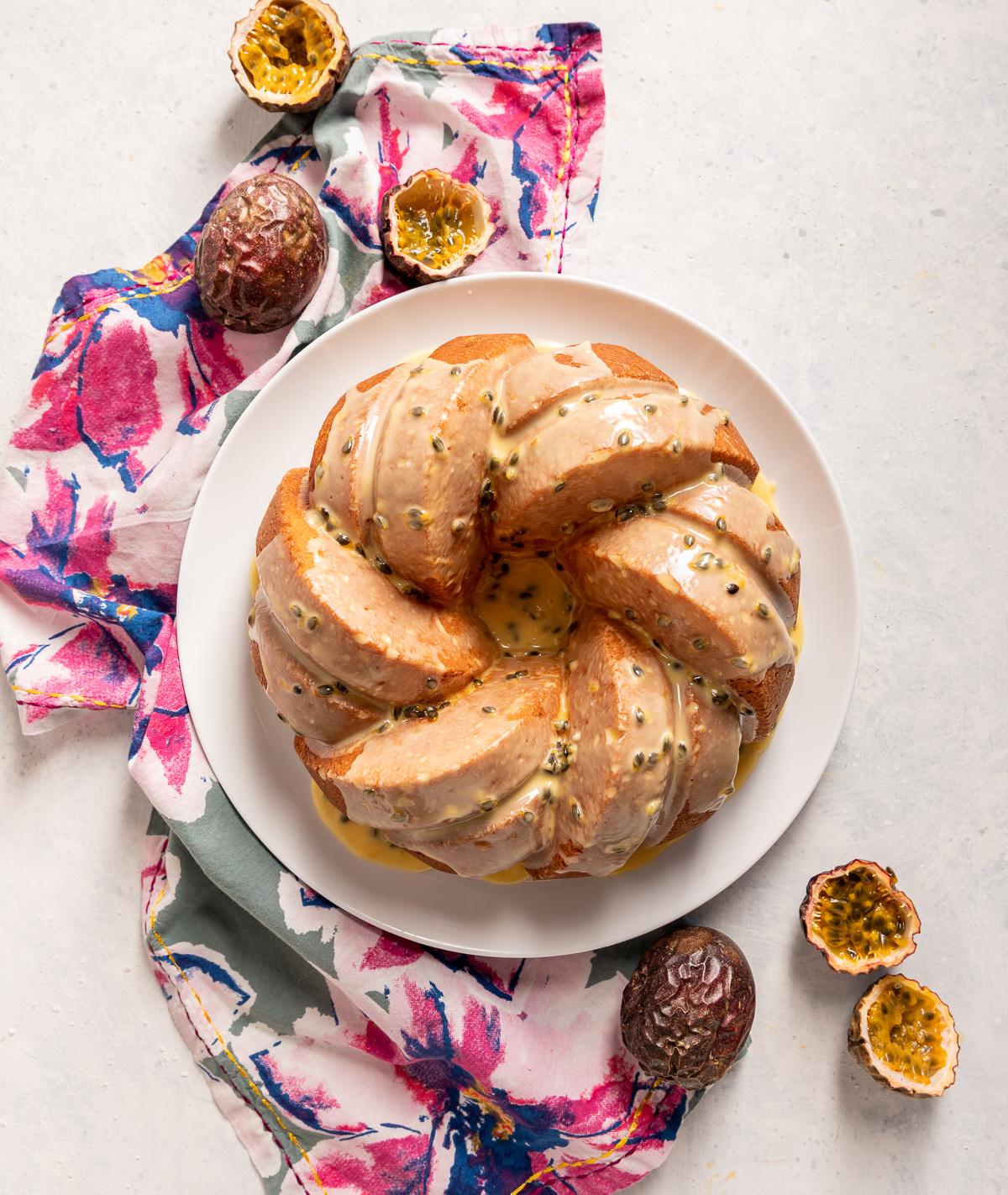 bundt cake with yellow passionfruit icing on white plate pink and blue flower napkin whole and halved passionfruits