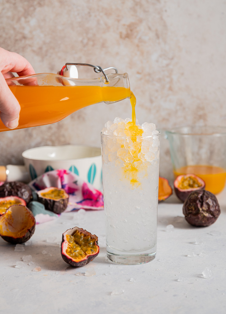 orange yellow syrup being poured into a glass filled with ice passion fruits cut in half
