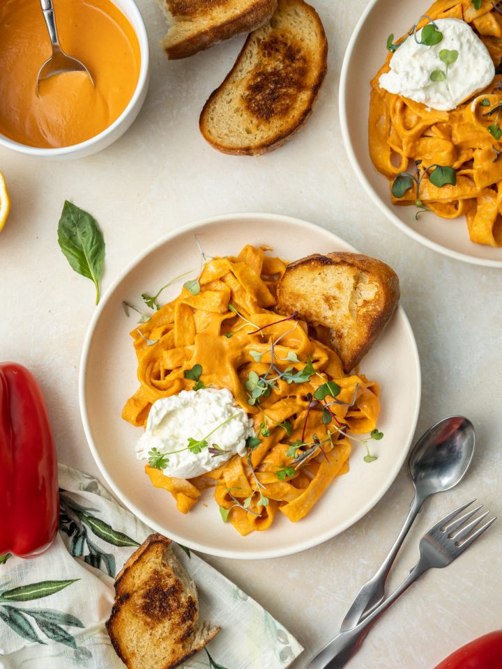 wide noodles with reddish orange roasted red pepper pasta sauce on a beige plate with slices of bread white burrata cheese and green sprouts and basil