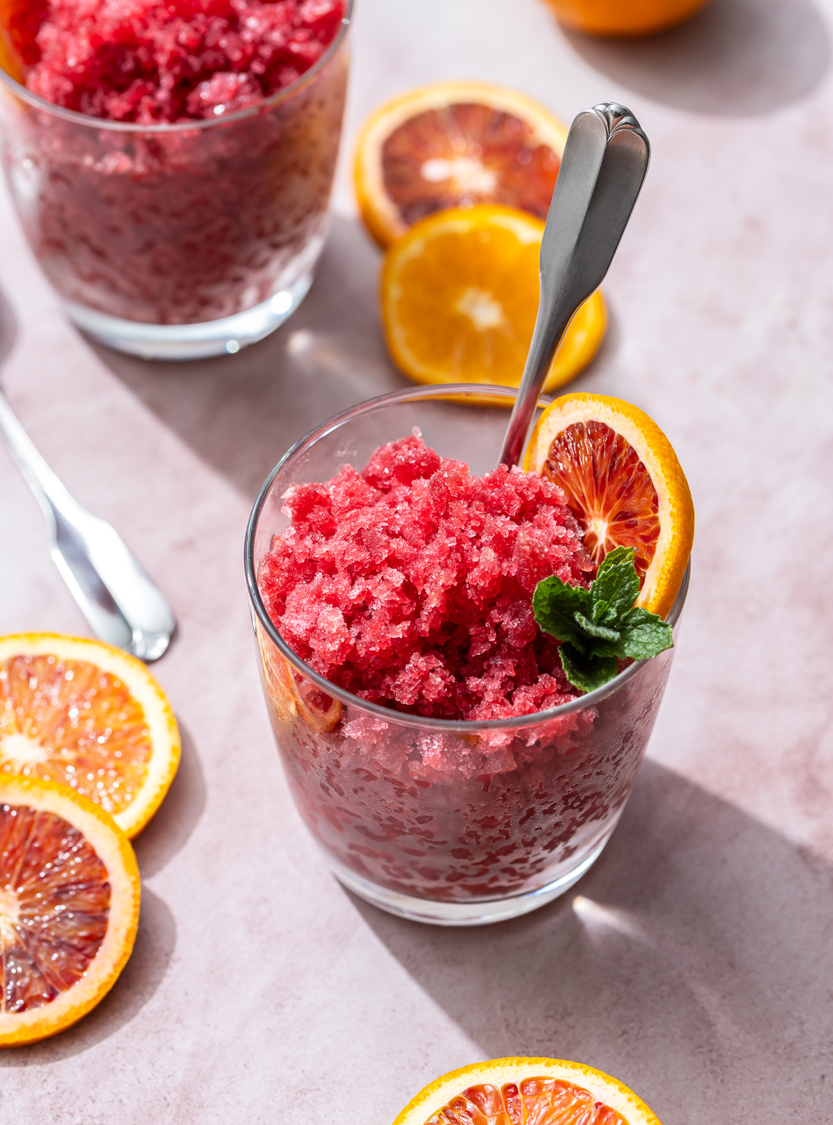 glass of reddish orange granita with mint blood orange and tangerine slices spoons