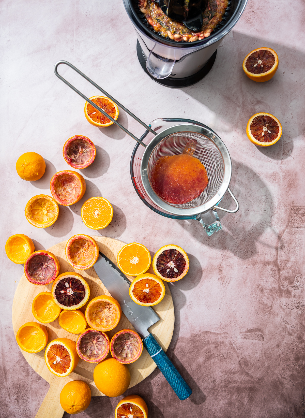 wood cutting board with blood oranges and tangerines cut in half strainer with citrus juice juicer and knife with blue handle