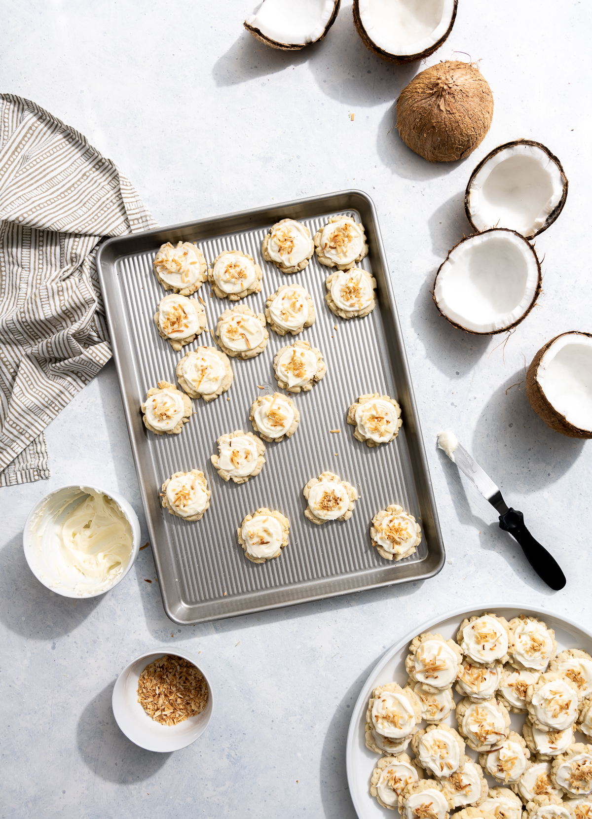 plate of coconut cookies bowl of coconut frosting bowl of toasted coconut fakes several cookies frosted spatula frosted cookies on a baking tray pieces of fresh coconut still in shell