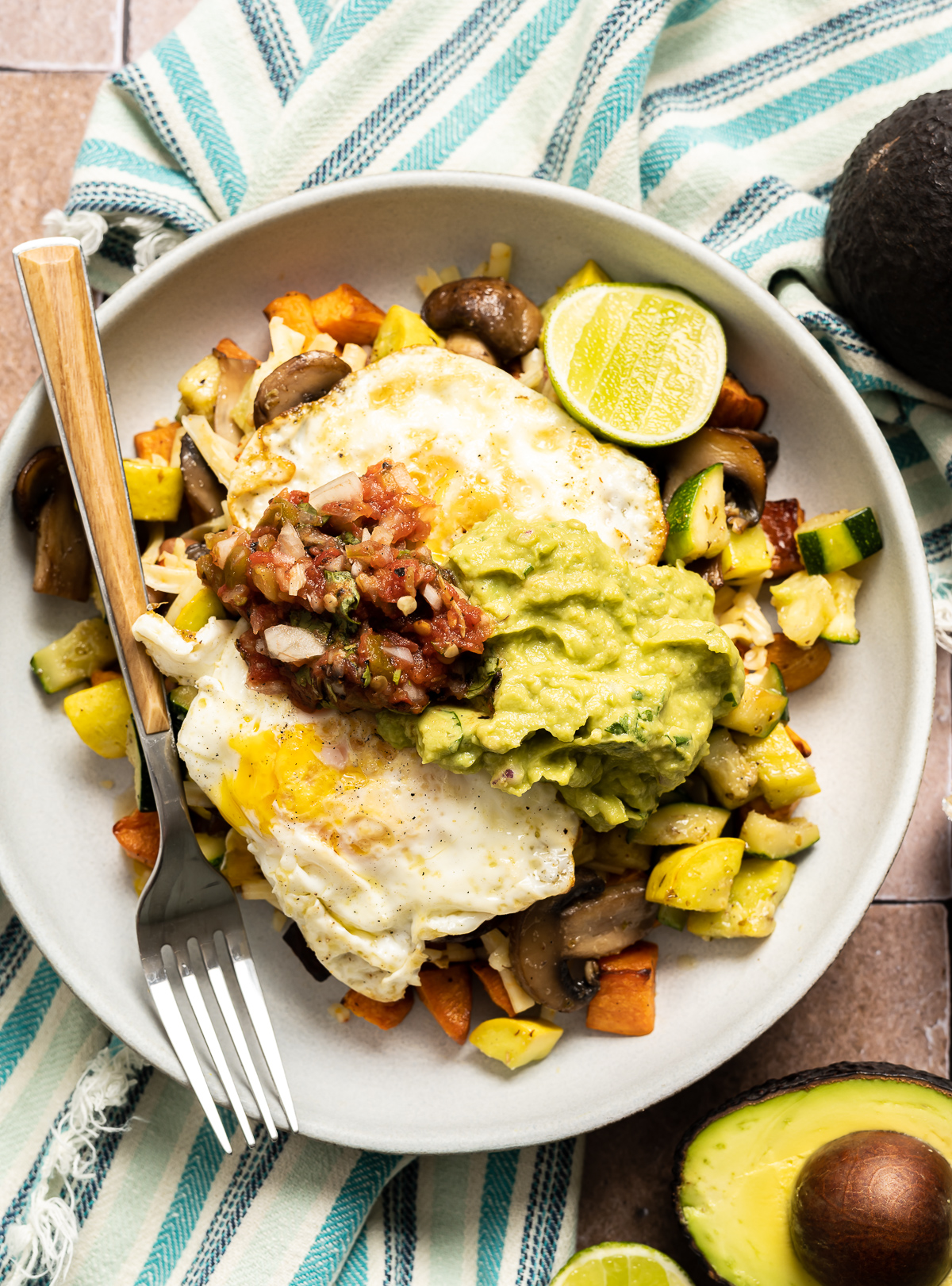bowl with diced zucchini squash sweet potato mushrooms with fried eggs spoonful of salsa and guacamole lime slices fork avocado halves