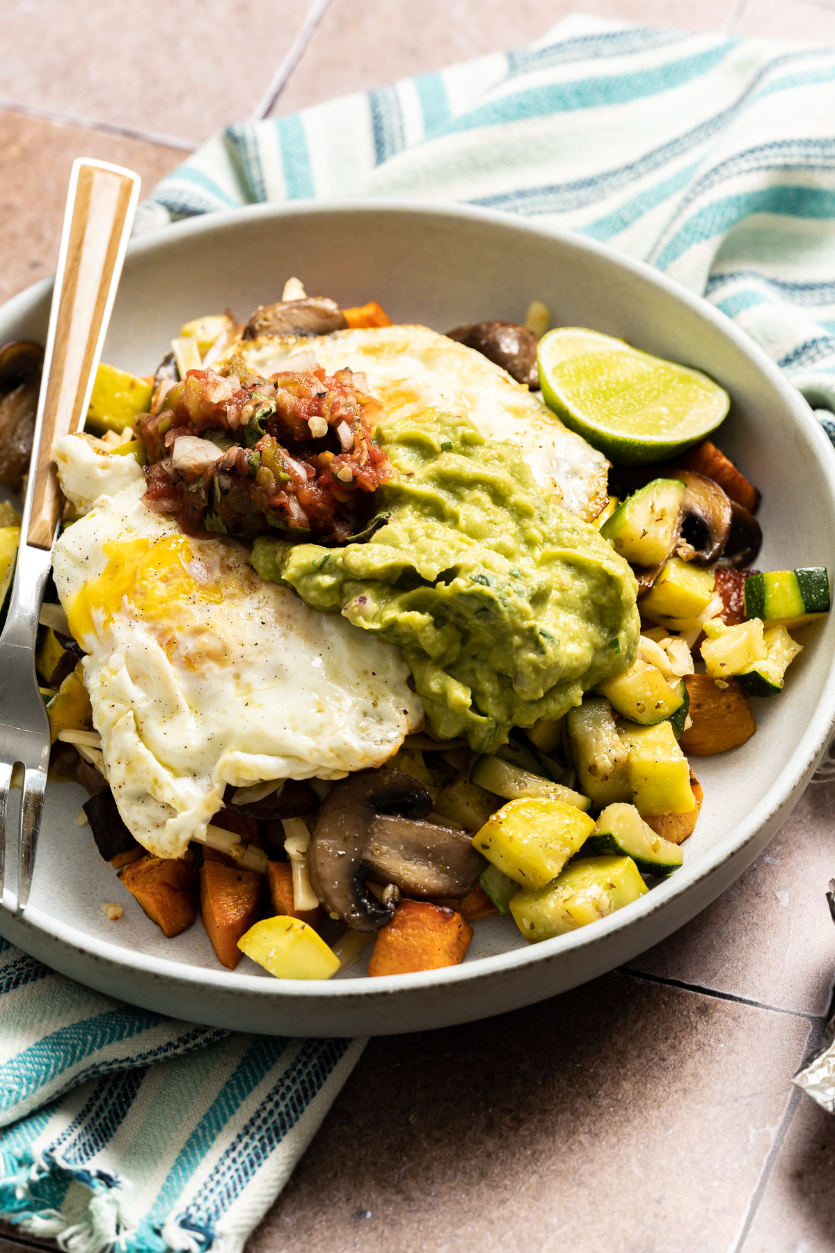 bowl with diced zucchini squash sweet potato mushrooms with fried eggs spoonful of salsa and guacamole lime slices fork avocado halves