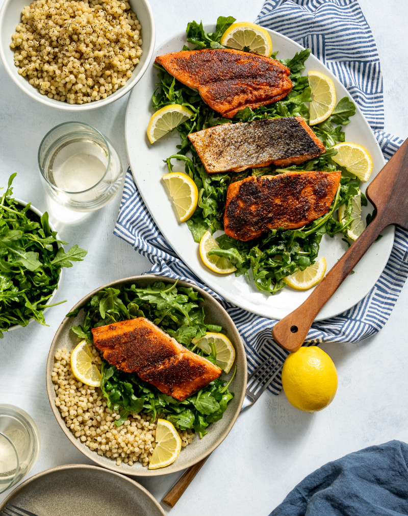 seared sockeye salmon on white and tan plates with green arugula yellow lemon slices bowl of israeli couscous