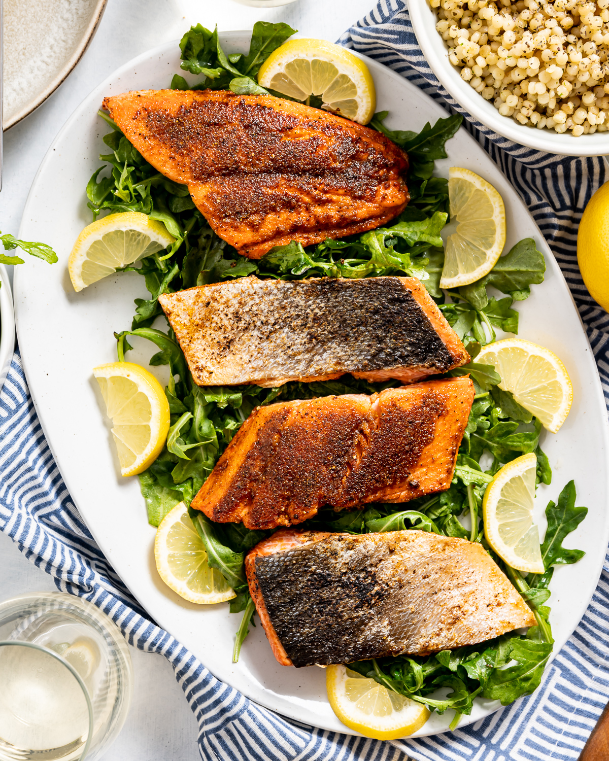 seared sockeye salmon on white plate with green arugula yellow lemon slices bowl of israeli couscous