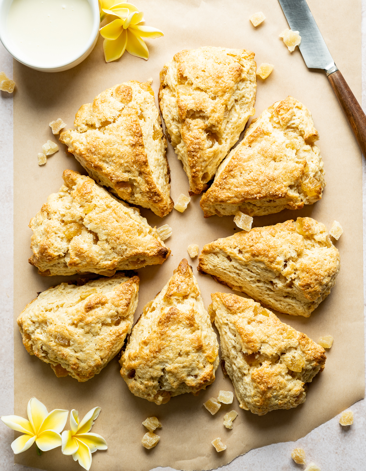 pineapple scones in a circle cut into wedges on brown parchment knife bowl of glaze yellow plumerias