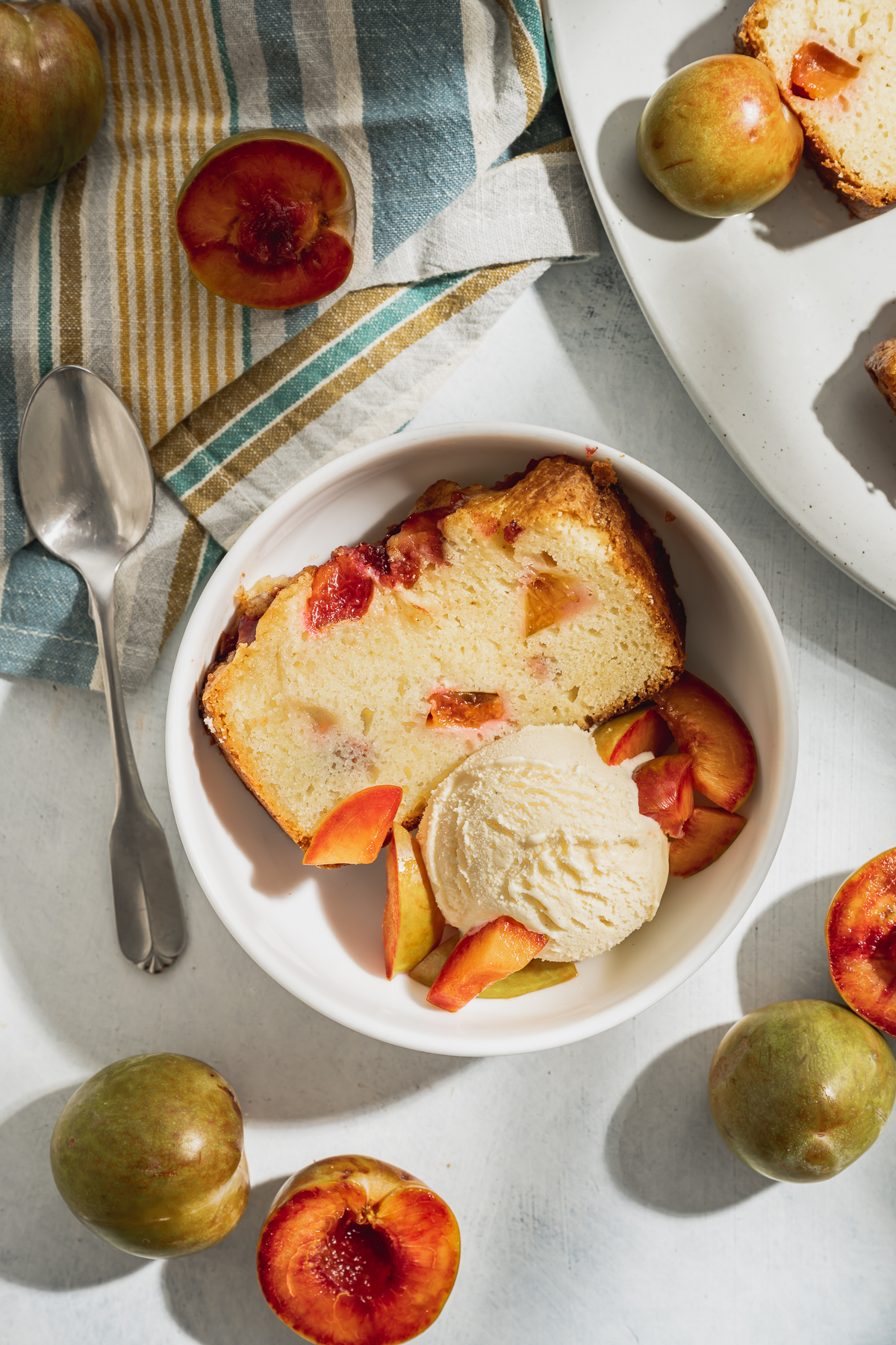 slice of plum pouncake in a bowl with a scoop of vanilla ice cream whole plums and sliced plums spoon striped napkin