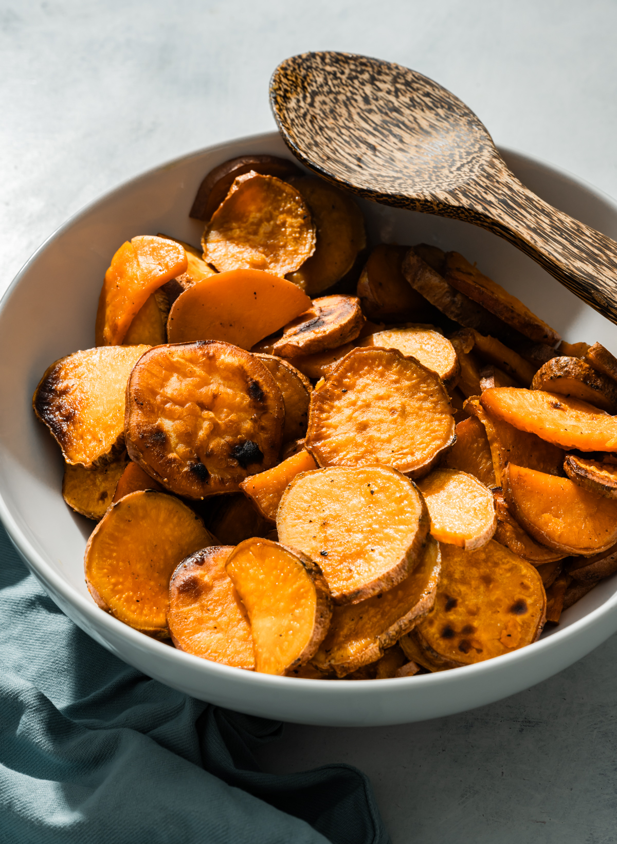 seasoned sliced roasted sweet potatoes in a white bowl wooden spoon blue napkin