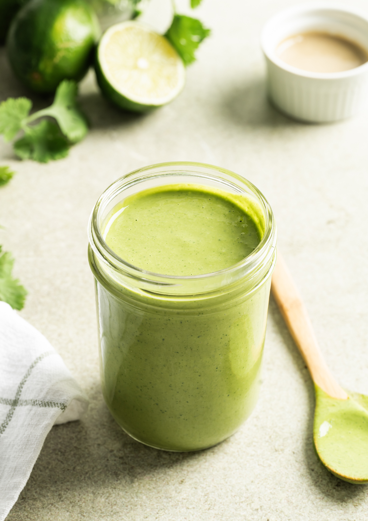 green dressing in a jar with wooden spoon next to it small bowl of tahini half a lime a few sprigs of cilantro
