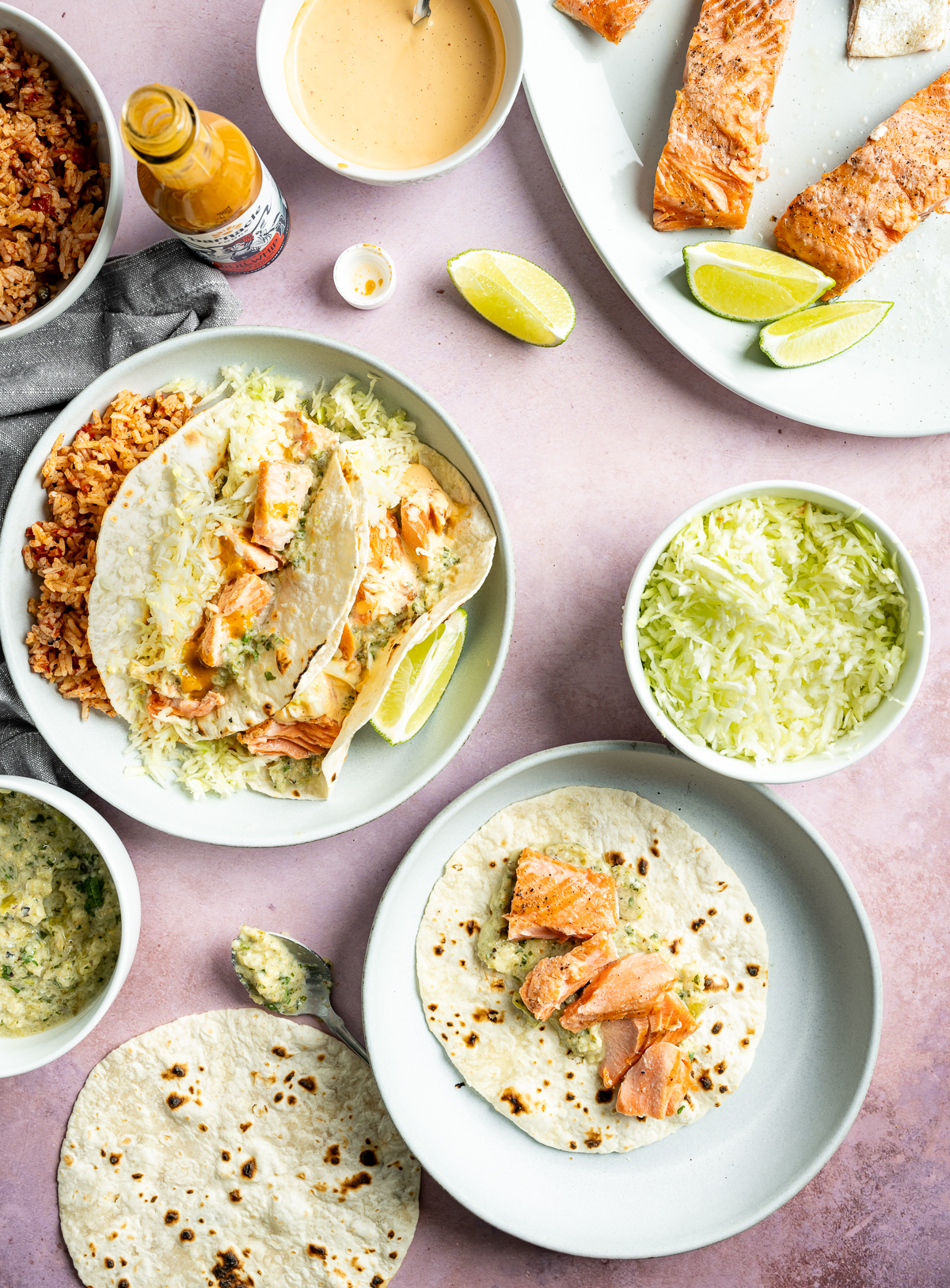 two plates of salmon tacos on light blue plates bowl of rice bowl of shredded green cabbage platter of grilled salmon
