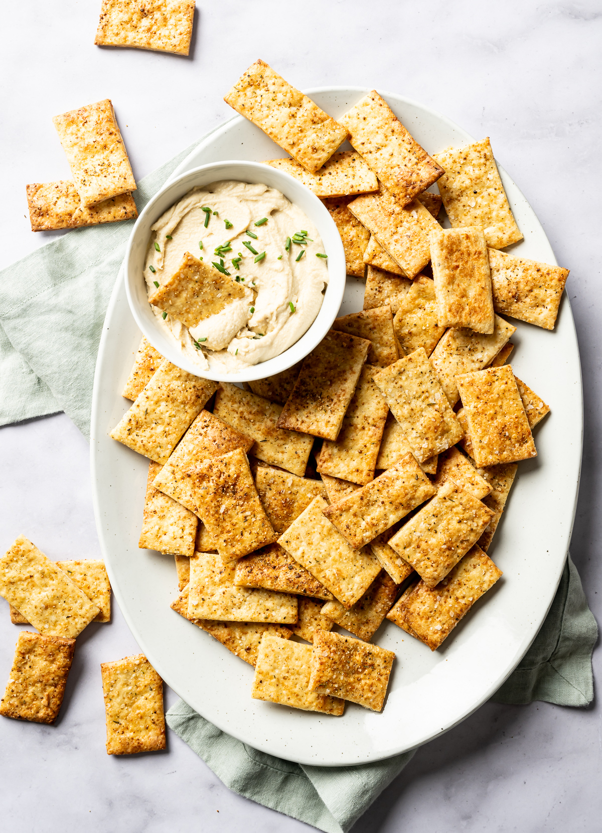 A white platter with a pile of golden baked cheese crackers with lemon pepper seasoning bowl of hummus with diced chives cracker in bowl of hummus