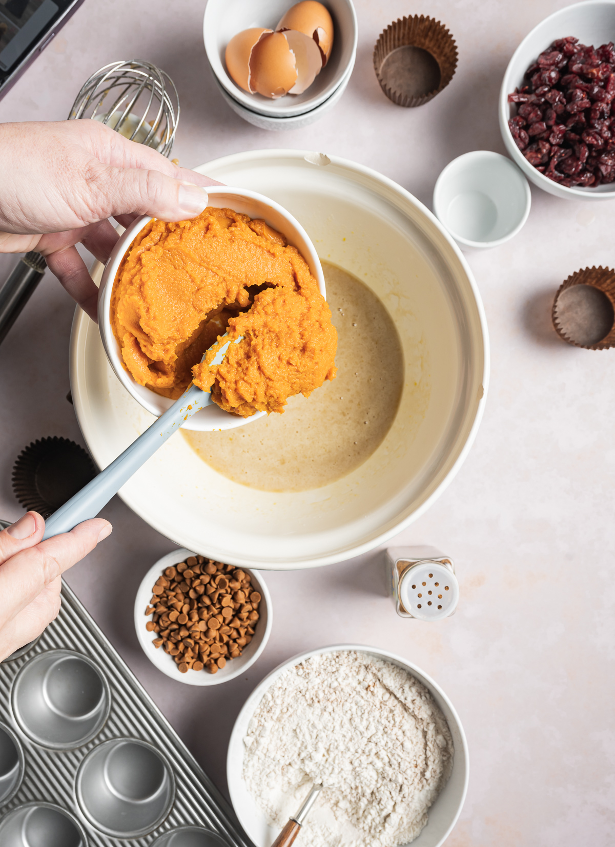 pumpkin puree being added to a bowl of muffin batter, various bowls of separate muffin ingredients