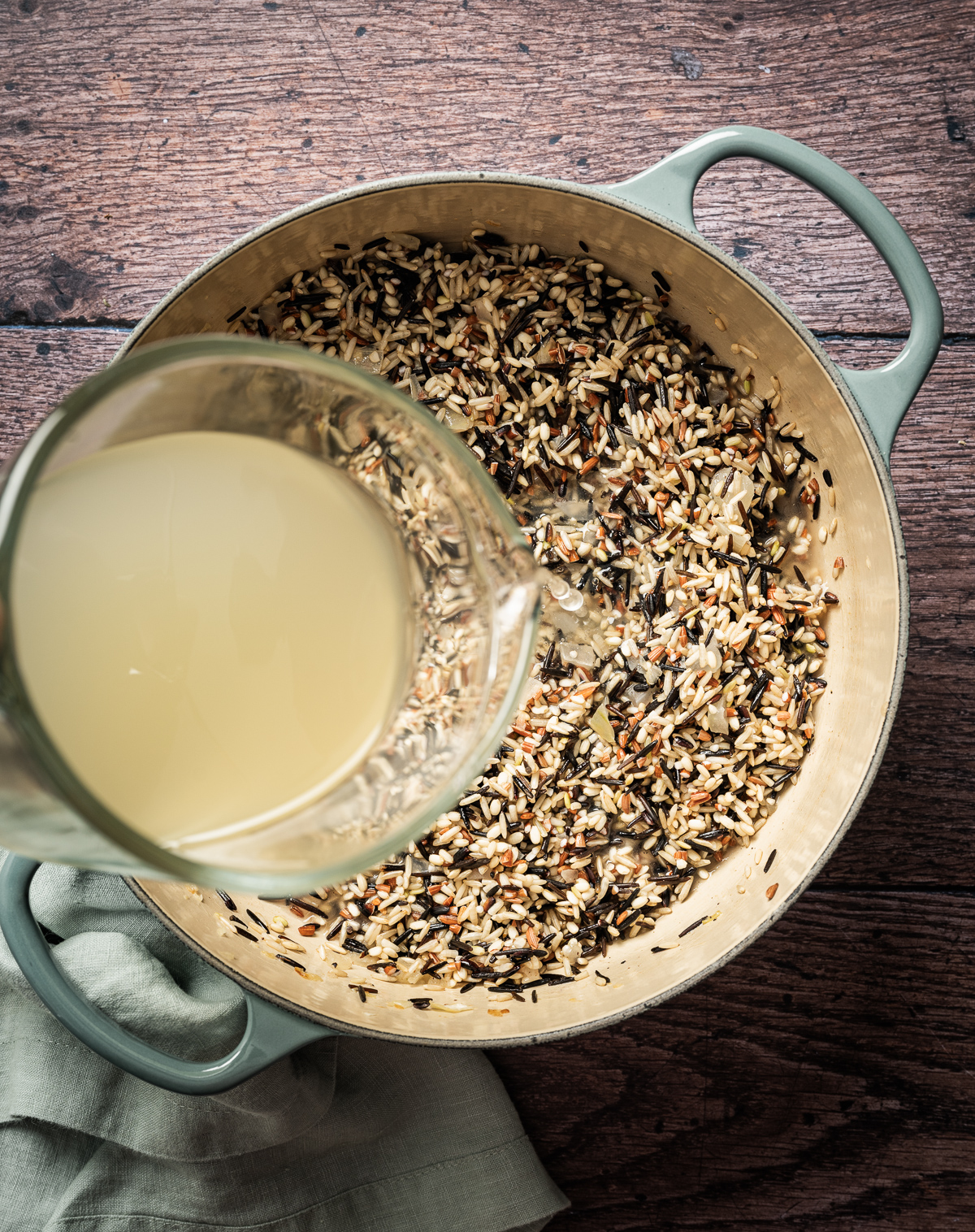 chicken broth being poured into large cast iron enamel pan with sauteed onion and wild rice