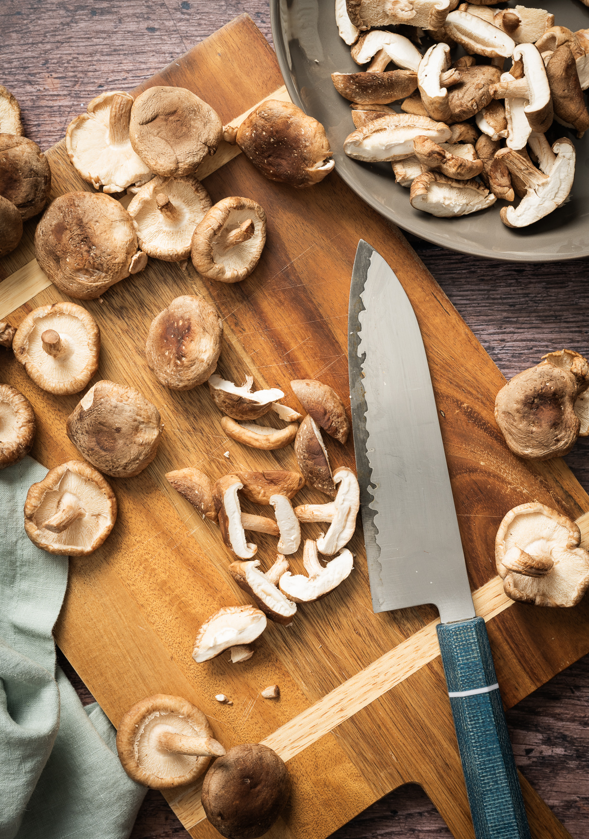 cutting board with whole and sliced fresh shiitake mushrooms blue handled japanese santoku knife